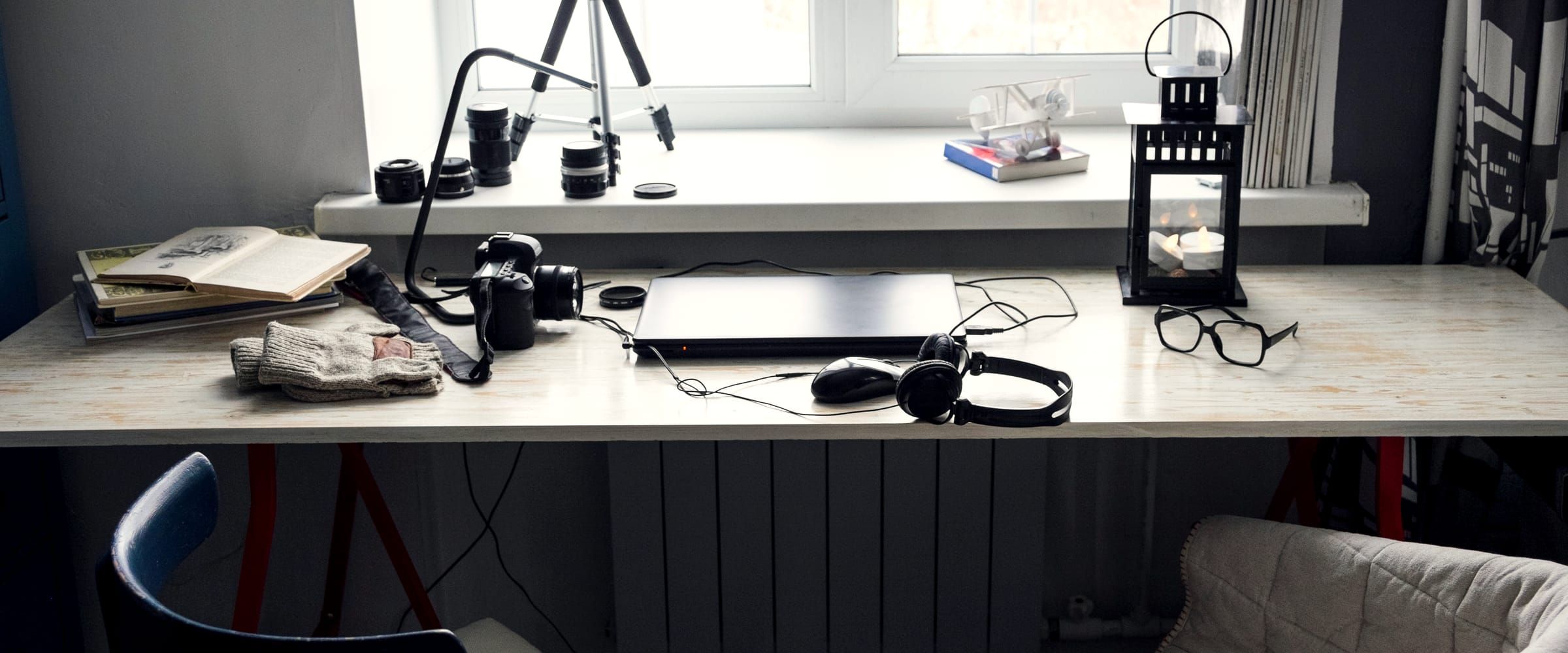 A workbench under a window holding a tripod, camera bodies, lenses and a laptop, with two chairs pulled up