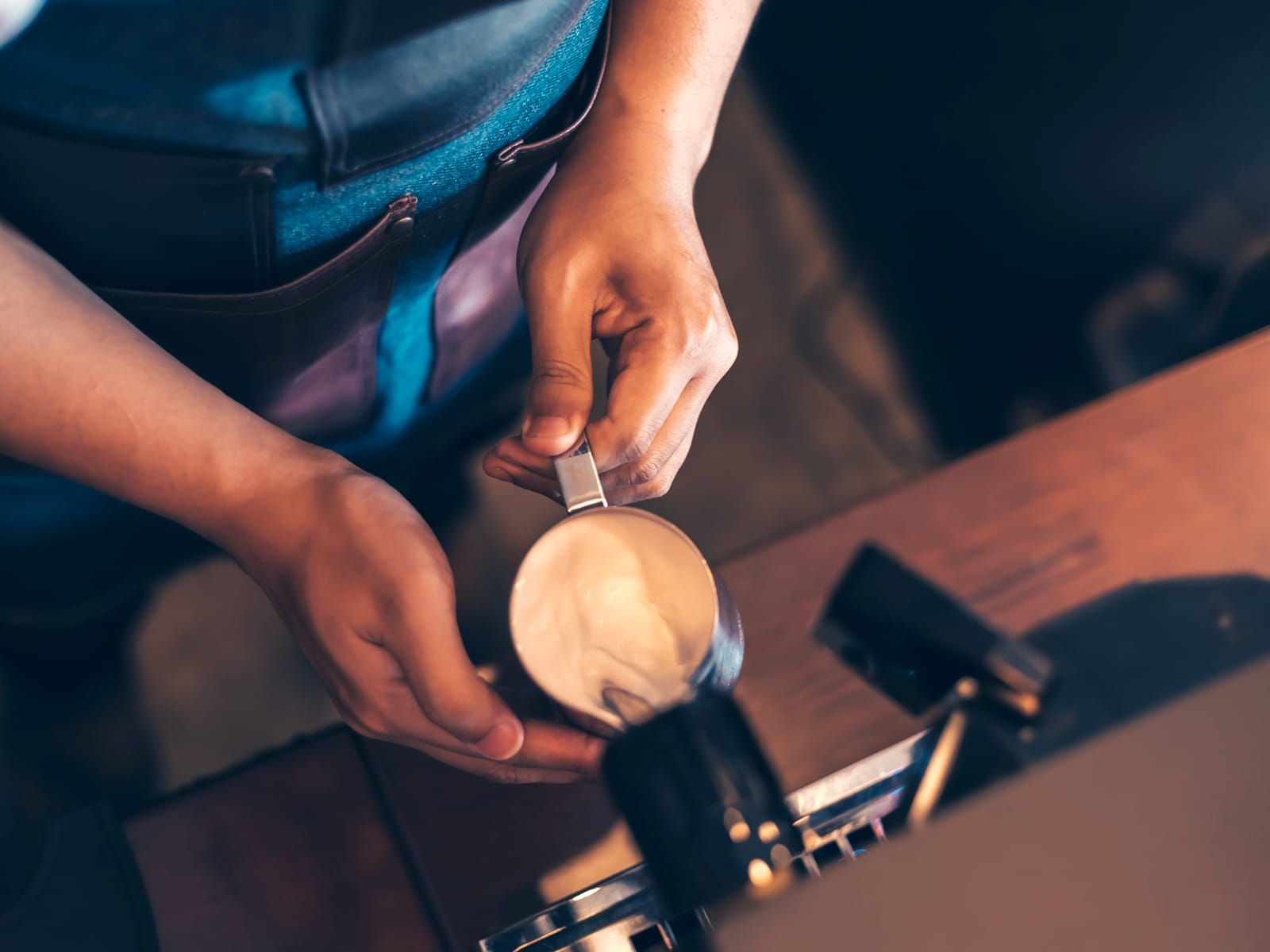 A barista’s hands frothing milk in a metal pitcher, seen from above