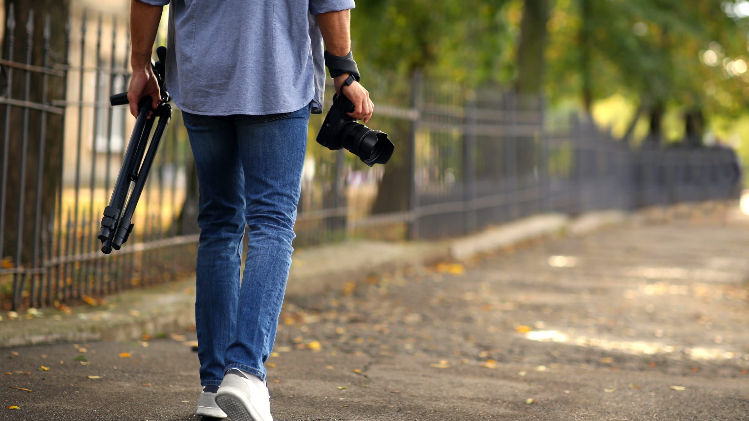 A photographer walking along a footpath, camera in one hand and folded tripod in the other